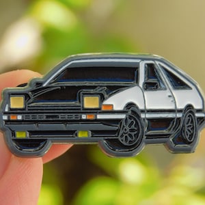 May include: A black and white enamel pin depicting a classic sports car. The car features a black roof and bonnet, a white body, and black wheels. The headlights and taillights are yellow and orange.