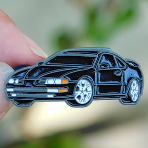 May include: Black car-shaped enamel pin with white and silver wheels, orange headlights, and a gray outline. The pin is held by a hand against a blurred green background. The car has a sleek, sporty design.