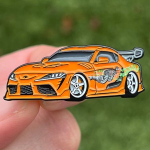 May include: Orange and black enamel pin of a Toyota Supra with a green and black graphic on the side. The car is facing the left side of the image.