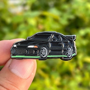 May include: Black enamel pin of a black car with a green outline. The car has a spoiler and is a popular model.