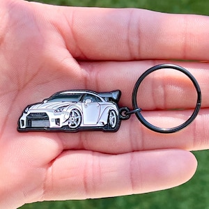 May include: A white and black car-shaped keychain with a black key ring. The car has detailed features, including wheels and a spoiler. The keychain is held in a hand against a blurred green background.