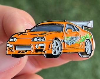 May include: Orange and black enamel pin of a Toyota Supra, a popular sports car, with a green and white design on the side. The pin is a popular collectible for fans of the Fast and Furious movie franchise.