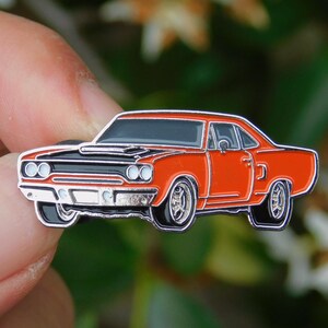 May include: A detailed enamel pin of a classic muscle car in orange and black. The pin features chrome detailing, black tyres, and a black bonnet. The car's body is a bright orange, and the pin has a glossy finish.