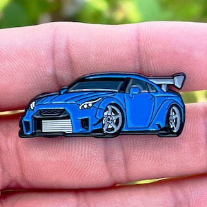 Könnte beinhalten: Eine blaue Emaille-Anstecknadel mit einem modifizierten Nissan Skyline GT-R Sportwagen mit einem abgedunkelten Fenster und einem Heckspoiler.