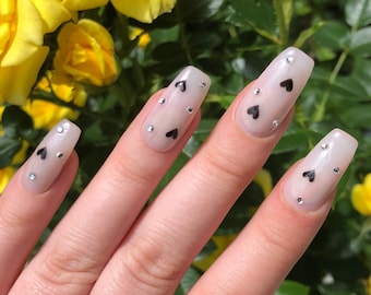 May include: Close-up of a hand with long, nude-colored nails with black heart designs and silver rhinestones. The hand is in front of a background of yellow roses.