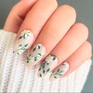 May include: Close-up of a hand with five nails painted with a light beige polish and a floral design. The design features white daisies with green leaves on a light beige background.