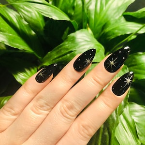 Puede incluir: Esmalte de uñas negro con diseños de estrellas plateadas. Las uñas son largas y en forma de almendra. La mano está frente a un fondo de hojas verdes.