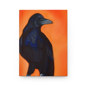 May include: Hardcover journal featuring a detailed illustration of a black crow against a vibrant orange background. The crow's feathers are depicted with intricate blue and black shading. The journal's cover is a solid white.