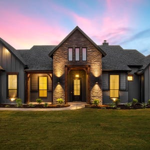 Può includere: Un moderno casale con facciata grigio scuro, dettagli in pietra e tetto grigio scuro. La casa presenta ampie finestre con calda illuminazione interna, sullo sfondo di un cielo colorato al tramonto e un prato ben curato.