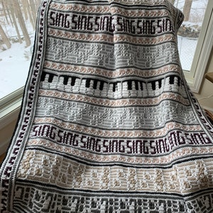 Puede incluir: Una manta de ganchillo hecha a mano en tonos blancos, grises, negros y beige. La manta presenta la palabra "SING" repetida en la parte superior y media, con teclas de piano y patrones geométricos. El borde es gris oscuro y negro.