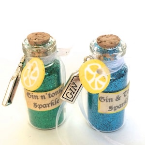 May include: Two small glass bottles filled with green and blue glitter. Each bottle has a cork stopper and a charm with a lemon slice and the word "GIN". The labels read "Gin n' tonic sparkle" and "Gin & tonic sparkle".