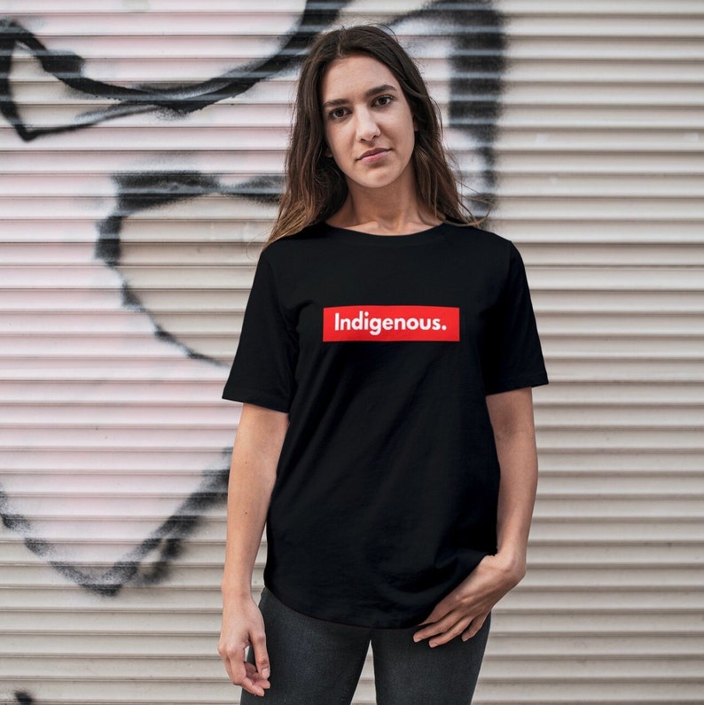 May include: Black t-shirt with the word "Indigenous" in white text inside a red rectangle. The t-shirt is a crew neck style and is being worn.