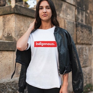 May include: A white t-shirt with the word "Indigenous" in a red box. The person is wearing a black leather jacket and black pants. The background is a stone wall.