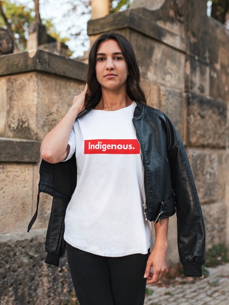 May include: A white t-shirt with the word "Indigenous" in a red box. The person is wearing a black leather jacket and black pants. The background is a stone wall.