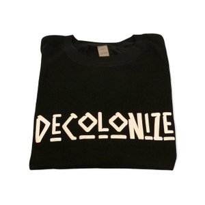 May include: Black crew neck t-shirt with the word "DECOLONIZE" printed in white block letters. The shirt is folded and appears to be made of a soft material. The text is centered on the front of the shirt.