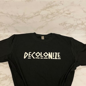 May include: Black t-shirt with the word "DECOLONIZE" printed in white, stylized block letters. The shirt is laid flat on a white and gray marble-patterned surface.