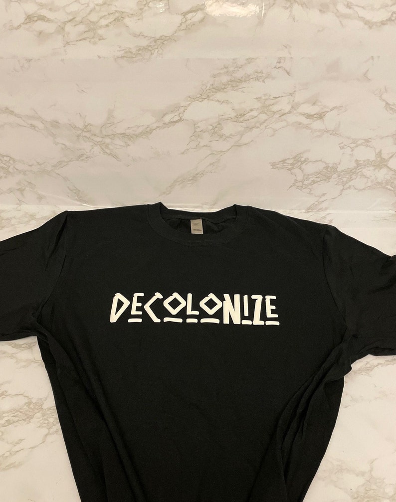 May include: Black t-shirt with the word "DECOLONIZE" printed in white, stylized block letters. The shirt is laid flat on a white and gray marble-patterned surface.