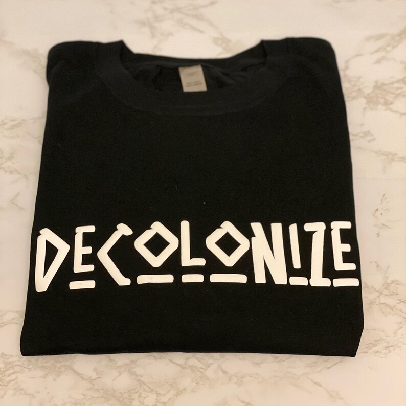May include: Black t-shirt with the word "DECOLONIZE" printed in white block letters. The shirt is folded neatly, showcasing the bold text. The design is simple and direct, making a statement.
