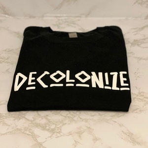 May include: A black t-shirt with the word "DECOLONIZE" printed in white, bold, stylized letters. The shirt is folded neatly, and the text is centered. The fabric appears to be a soft material.