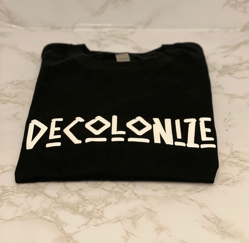 May include: A black t-shirt with the word "DECOLONIZE" printed in white, bold, stylized letters. The shirt is folded neatly, and the text is centered. The fabric appears to be a soft material.