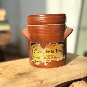 May include: A brown ceramic jar with two handles and a label that reads "Moutarde de Luxe". The jar is cylindrical with a lid and sits on a wooden surface. The label is yellow and black with red text.