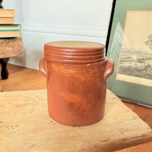May include: A brown ceramic jar with a lid, featuring two small handles and a textured finish. The jar is placed on a wooden surface. This item is likely used for storage or decorative purposes.