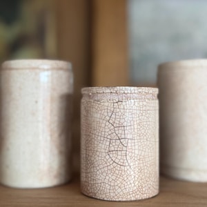May include: Three cylindrical ceramic vessels with a cream-coloured glaze featuring a prominent dark brown crackle pattern. The vessels have a straight-sided shape with a slightly raised rim at the top, displaying a uniform texture and finish.