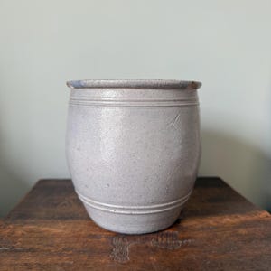 May include: A gray stoneware crock with a cylindrical shape. The antique jar has a slightly flared rim and two horizontal bands around the body. A single vertical mark is visible on the side. The crock sits on a wooden surface.