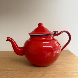 May include: A vibrant red enamel teapot with a curved spout and a matching lid. The teapot has a rounded body and a black trim around the lid. The handle is a curved metal loop. The teapot is sitting on a wooden surface.