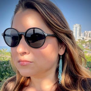 Può includere: Una donna con occhiali da sole rotondi neri e una maglietta nera con lunghi orecchini a perline blu.