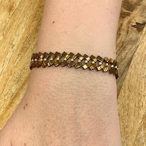 May include: A gold bracelet with a geometric design and a gold ring on a finger with a light pink nail polish.