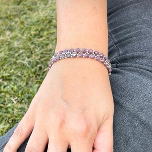 Könnte beinhalten: Nahaufnahme einer Hand, die ein doppelreihiges Armband mit violetten Perlen trägt. Das Armband besteht aus kleinen, runden Perlen, die auf einem silbernen Draht aufgereiht sind.