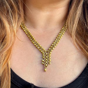 Puede incluir: Un collar de color dorado con un diseño en forma de V y un colgante en forma de lágrima. El collar está hecho de pequeñas cuentas redondas.