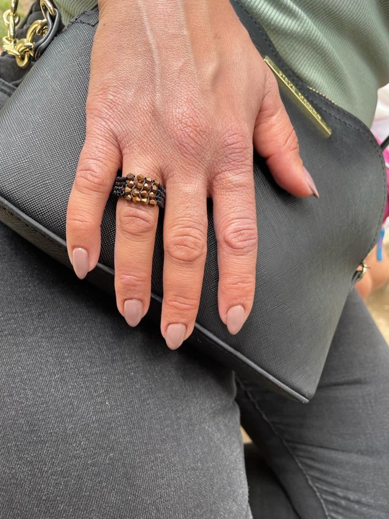 K&ouml;nnte beinhalten: Nahaufnahme einer Hand, die einen goldenen und schwarzen Perlenring tr&auml;gt. Der Ring besteht aus kleinen, runden Perlen, die in einem geometrischen Muster angeordnet sind. Die Hand ruht auf einer schwarzen Lederhandtasche.