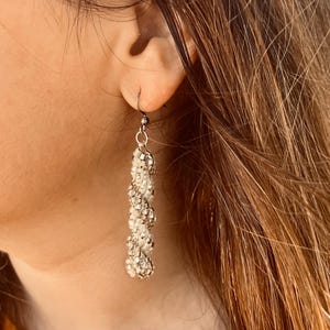 May include: Silver dangle earrings with a beaded design. The earrings have a long, vertical strand of alternating silver and white beads, creating a textured accessory. The earrings are suspended from a simple hook. A stylish accessory.