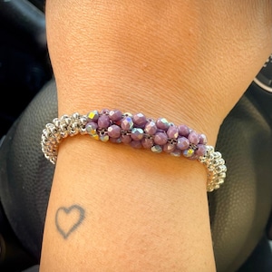 Pode incluir: Um bracelete de contas prateadas e roxas com uma tatuagem de coração no pulso.