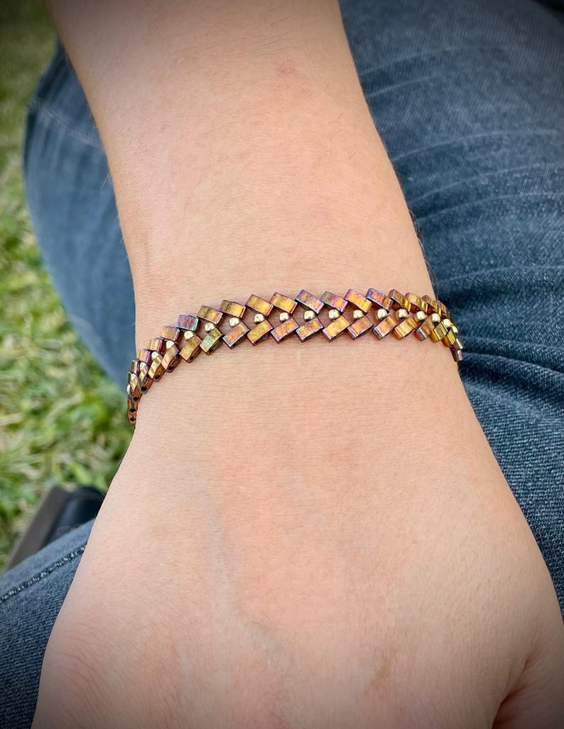 May include: A woven bracelet with a geometric pattern in shades of brown, gold, and purple. The bracelet is made of small, square-shaped beads.