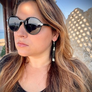 May include: A pair of long, black and white beaded earrings. The earrings are hanging from the woman's ears. The woman is wearing black sunglasses and a black shirt.