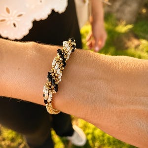 Könnte beinhalten: Ein Armband mit Perlen, das eine goldene Basis hat und mit schwarzen, weißen und goldenen Perlen verziert ist. Das Armband wird am Handgelenk getragen. Ein weißes Oberteil und eine schwarze Hose sind im Hintergrund zu sehen.
