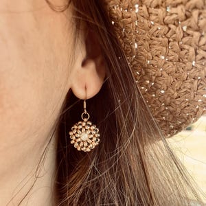 May include: Gold-colored beaded earrings with a floral design. The earrings have a central white bead surrounded by smaller gold beads. The earrings are hanging from a gold-colored hook. The background includes a straw hat and hair.