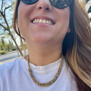 May include: A close-up of a woman wearing a gold and purple beaded necklace. The necklace is made of small, round beads that are arranged in a geometric pattern. The woman is smiling and wearing sunglasses.