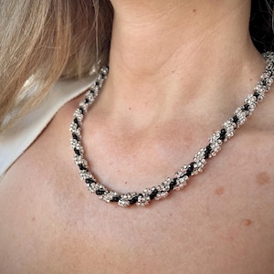 Op de afbeelding: Een zilver-zwarte kralenketting. De ketting is gemaakt van afwisselende rijen zilverkleurige kralen en zwarte kralen, waardoor een gedraaid touwontwerp ontstaat. De ketting wordt om de nek gedragen.
