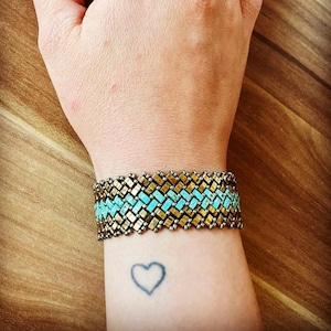 Op de afbeelding: Een turquoise en gouden kralen armband met een chevron patroon. De armband wordt gedragen op iemands pols.