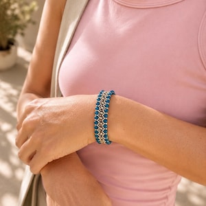 Puede incluir: Un brazalete de cuentas turquesas y doradas. El brazalete se lleva en la muñeca, sobre una camiseta rosa. El diseño presenta filas de pequeñas cuentas redondas turquesas separadas por cuentas doradas. Joyería.