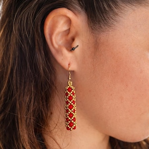 Puede incluir: Pendientes de cuentas doradas con un diseño rectangular. Los pendientes presentan filas alternas de pequeñas cuentas redondas rojas y doradas. Los pendientes cuelgan de un gancho dorado. La imagen muestra los pendientes en una oreja.