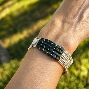 May include: A gold-toned bracelet with a central band of dark green, square beads. The bracelet features multiple strands of small, round gold beads, creating a woven effect. The bracelet is worn on a wrist against a blurred green background.