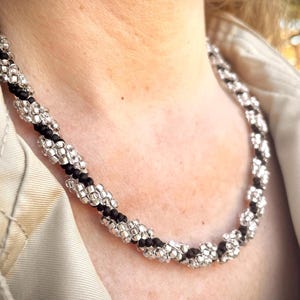 Może przedstawiać: Naszyjnik z czarnych i srebrnych koralików o skręconym wzorze. Naszyjnik wykonany jest z małych, okrągłych koralików, które są nawleczone w spiralny wzór.