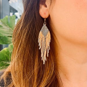 Può includere: Orecchini con perline color oro con una sezione superiore con perline grigie e beige e lunghe ciocche pendenti. Gli orecchini sono a forma di goccia e vengono indossati sull'orecchio di una persona.