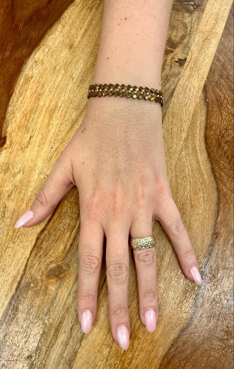 May include: A gold bracelet with a geometric design and a gold ring on a finger with a light pink nail polish.