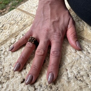 K&ouml;nnte beinhalten: Eine Hand tr&auml;gt einen goldenen Ring mit mehreren Reihen kleiner, dunkelbrauner Steine. Die N&auml;gel sind in einem hellen Braunton lackiert.
